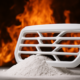 A white automotive grille-shaped engineering plastic component is placed atop a mound of white pigment powder, with flames blazing in the background to symbolize extreme heat. The image visually highlights the heat resistance and color stability of GEOLOR® ST high-performance pigment.